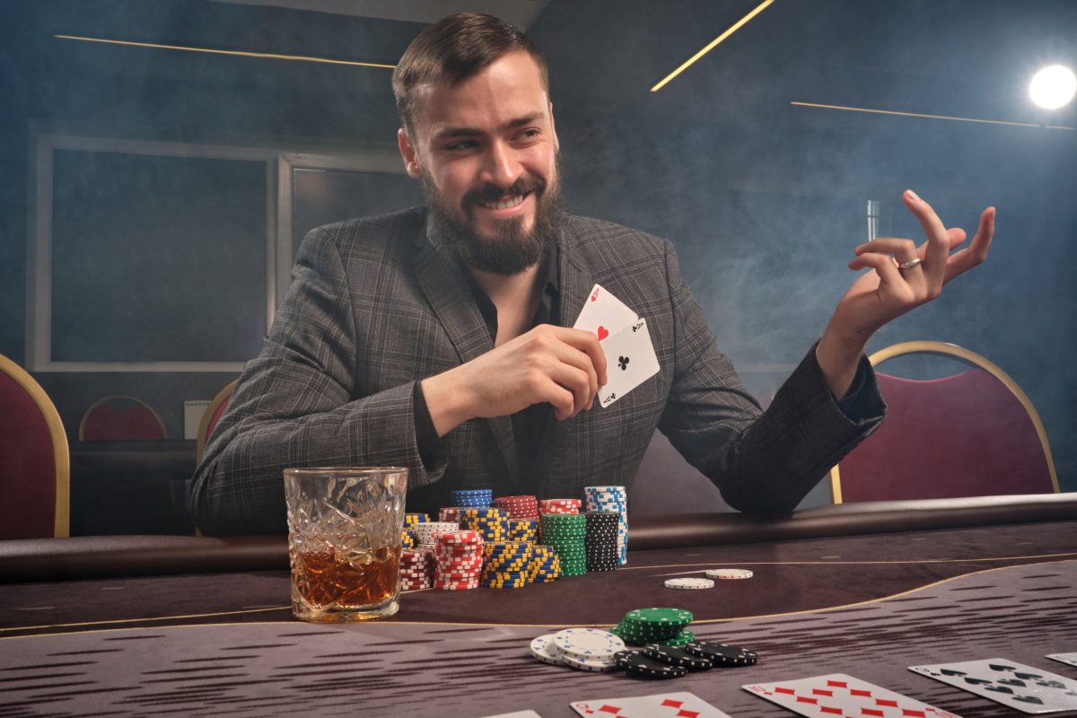 Cheeky brunette man in a classic suit is playing poker sitting at the table at casino in a smoke. He is making bets waiting for a big win. Gambling for money. Games of fortune.